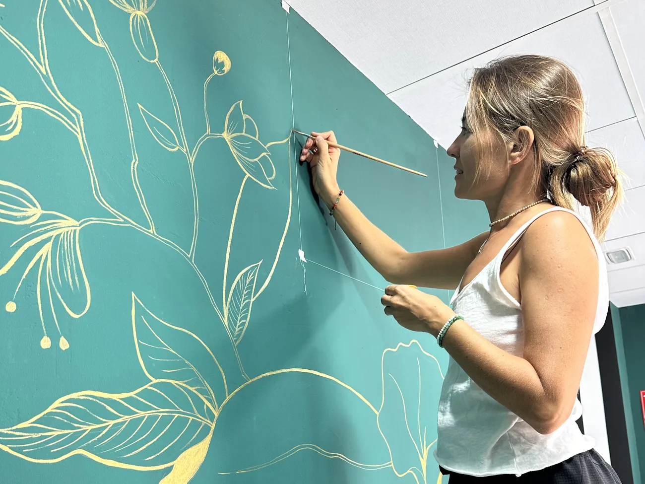 María pintando a mano un mural floral dorado sobre una pared verde, en un interior