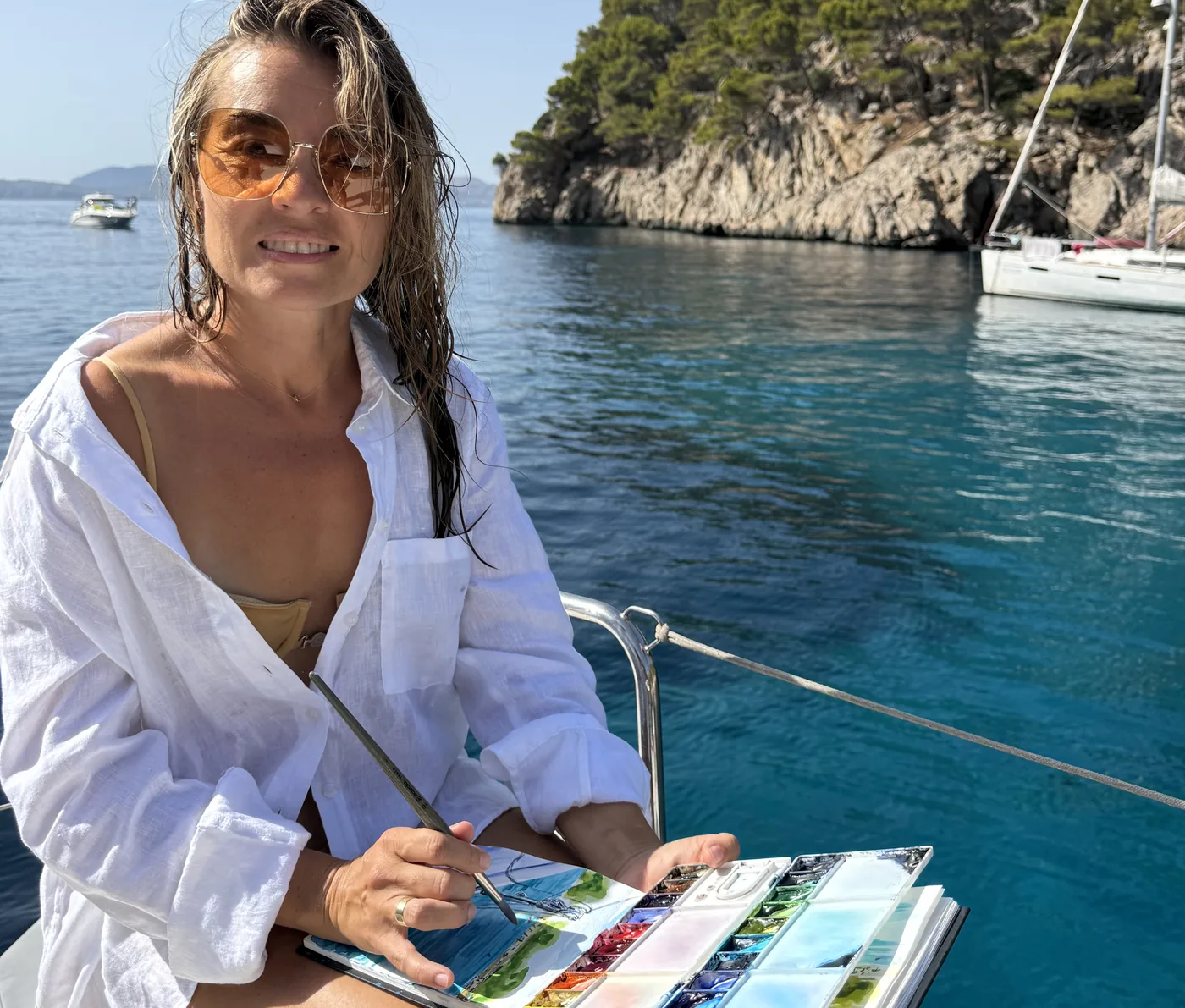 María sonríe con la paleta de acuarelas y el cuaderno abiertos, pintando al aire libre junto al mar